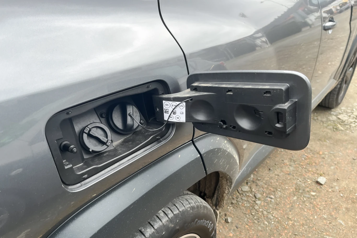 Otwarta klapka wlewu paliwa w szarym samochodzie terenowym, widoczna instrukcja obsługi oraz korek zabezpieczający otwór wlewu. Tło stanowią szutrowa nawierzchnia i fragment tylnego nadkola pojazdu.