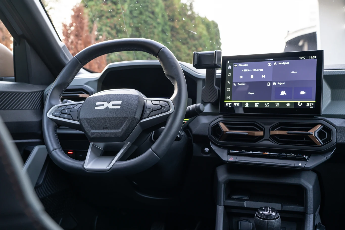 Nowoczesna deska rozdzielcza samochodu z wyświetlaczem dotykowym, kierownicą z logo producenta oraz elektronicznymi wskaźnikami. Po lewej stronie widoczna część drzwi, z przodu osłona silnika, a w tle zieleń i fragment białej karoserii.