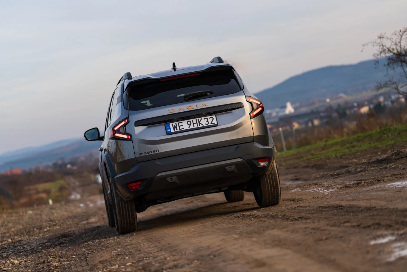 Szary samochód marki Dacia Duster porusza się po nieutwardzonej drodze na tle łagodnych wzgórz i rozległego krajobrazu, w oddali widoczne pojedyncze zabudowania. Centralnym elementem jest tył pojazdu z czytelną tablicą rejestracyjną.