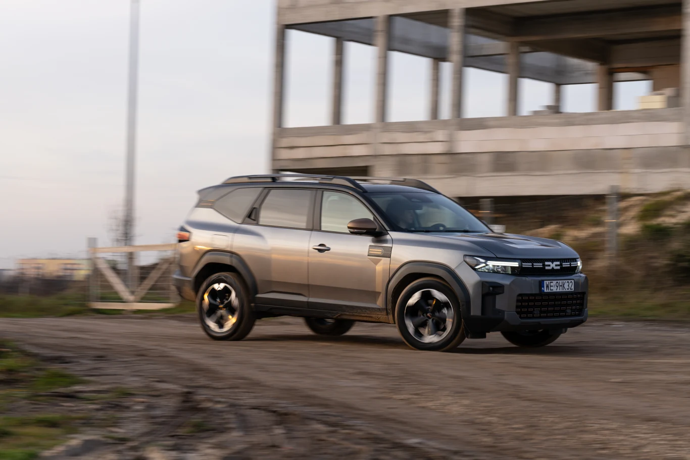 Nowoczesny SUV w kolorze szarym jedzie po nieutwardzonej drodze obok niedokończonego betonowego budynku, w tle widoczne są elementy miejskiego krajobrazu.