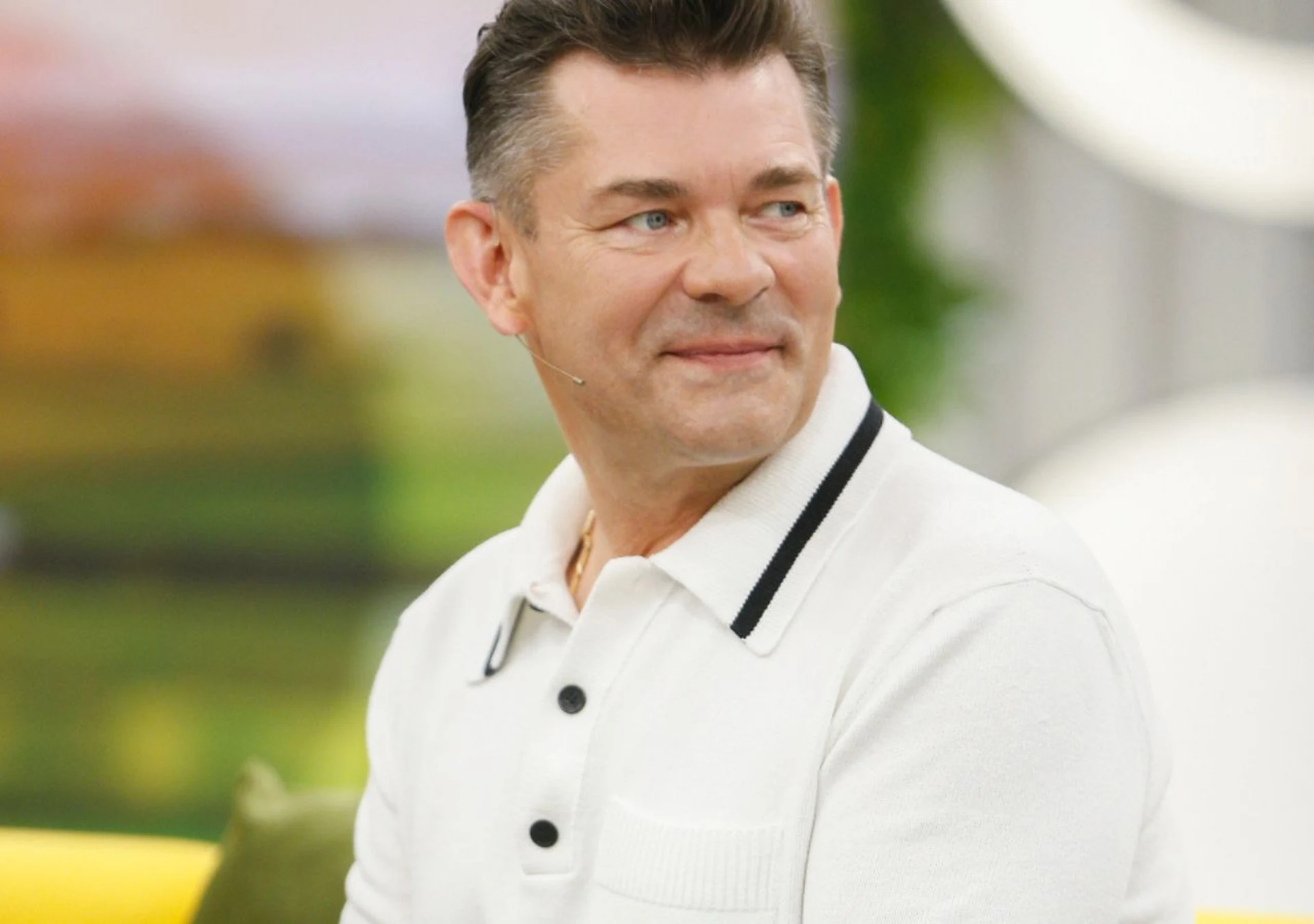 Mężczyzna o krótkich, ciemnych włosach w białej koszulce polo z czarnymi akcentami, siedzący na tle rozmytego, zielonego wnętrza, z delikatnym uśmiechem na twarzy.