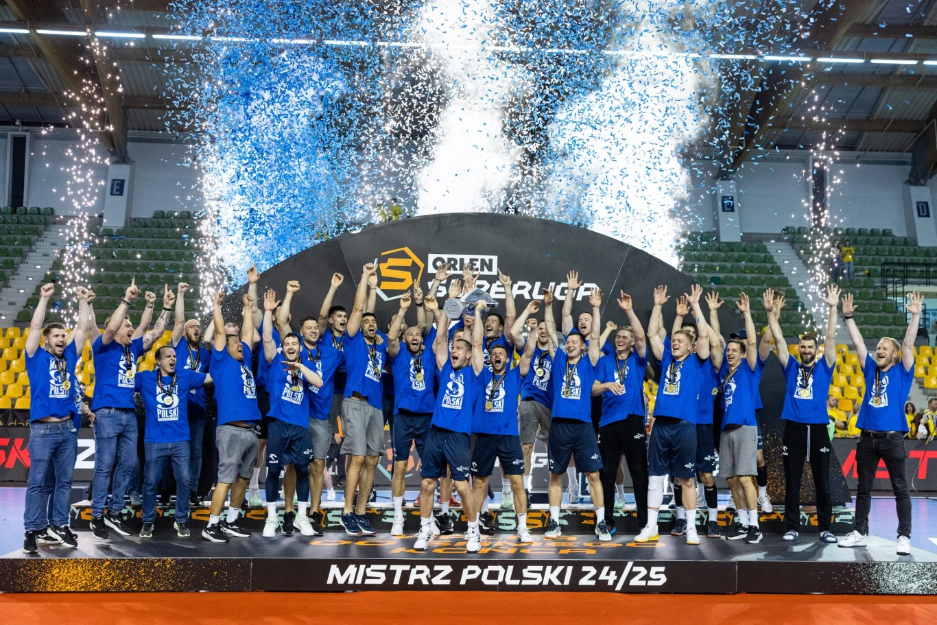 Drużyna mężczyzn w niebieskich koszulkach świętuje zdobycie tytułu mistrza Polski, stojąc na podium z medalami na szyi, uniesionymi rękami i konfetti sypiącym się z góry, w tle widoczne efekty specjalne i napisy związane z wydarzeniem sportowym.