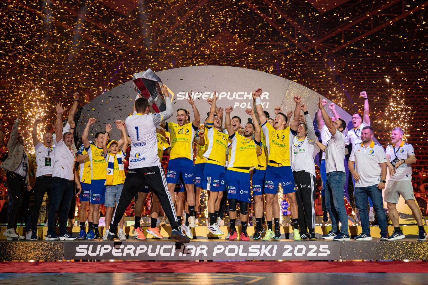 Drużyna piłkarzy ręcznych w żółto-niebieskich strojach świętuje zdobycie Superpucharu Polski 2025, unosi trofeum w otoczeniu sztucznych ogni i konfetti na scenie z napisem konkursowym, wokół nich stoją trenerzy oraz członkowie sztabu.