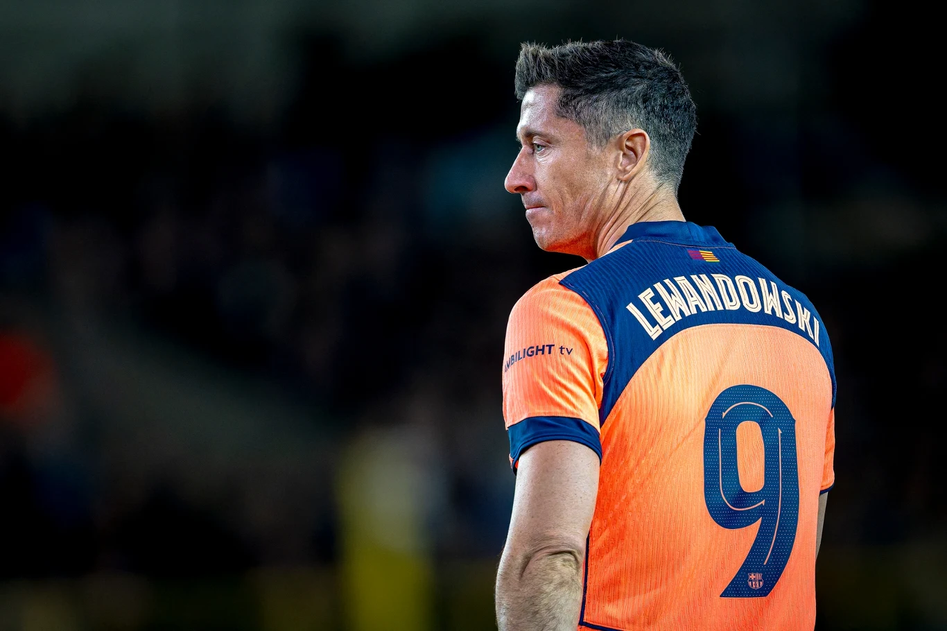 Piłkarz ubrany w pomarańczową koszulkę z numerem 9 i nazwiskiem Lewandowski na plecach, patrzący w bok, na tle rozmazanej, ciemnej trybuny stadionu.