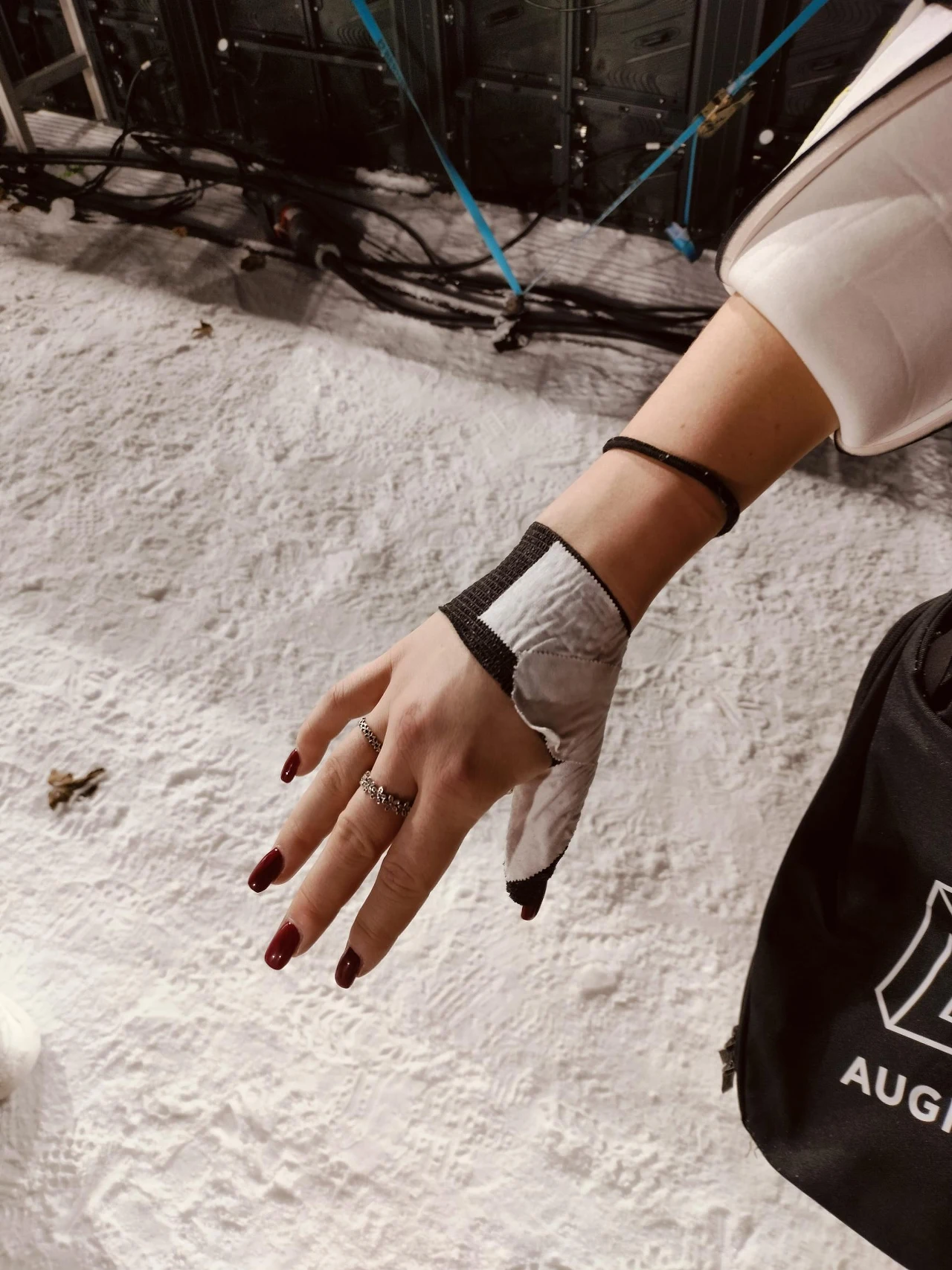 Ręka kobiety z paznokciami pomalowanymi na ciemnoczerwono, częściowo owinięta taśmą sportową lub opaską stabilizującą w nadgarstku, widoczna na tle jasnej, teksturowanej powierzchni. W tle czarny plecak z logo oraz przewody techniczne.