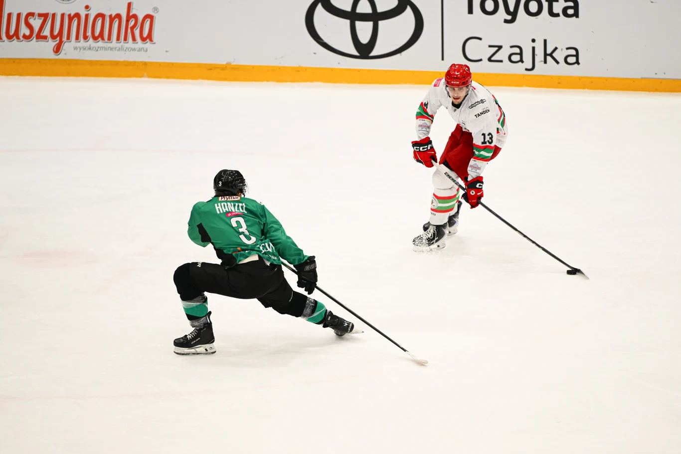 Dwóch hokeistów na lodzie, ubranych w kolorowe stroje, uczestniczy w dynamicznej akcji meczowej. Jeden z zawodników jest w pozycji obronnej, pochylony i przygotowany do zablokowania przeciwnika, podczas gdy drugi prowadzi krążek hokejowy. Na bandzie wi...