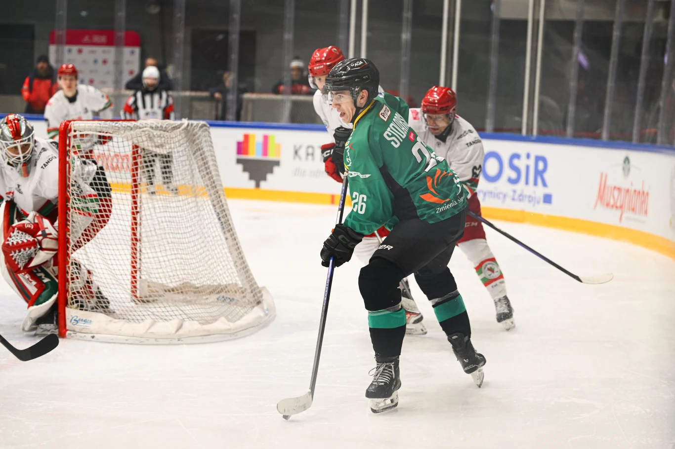 Zawodnicy drużyn hokejowych rywalizują na lodowisku, skupieni wokół bramki, jeden z graczy w zielonym stroju próbuje zdobyć gola, podczas gdy bramkarz oraz obrońcy drużyny przeciwnej w białych i czerwonych strojach koncentrują się na obronie.