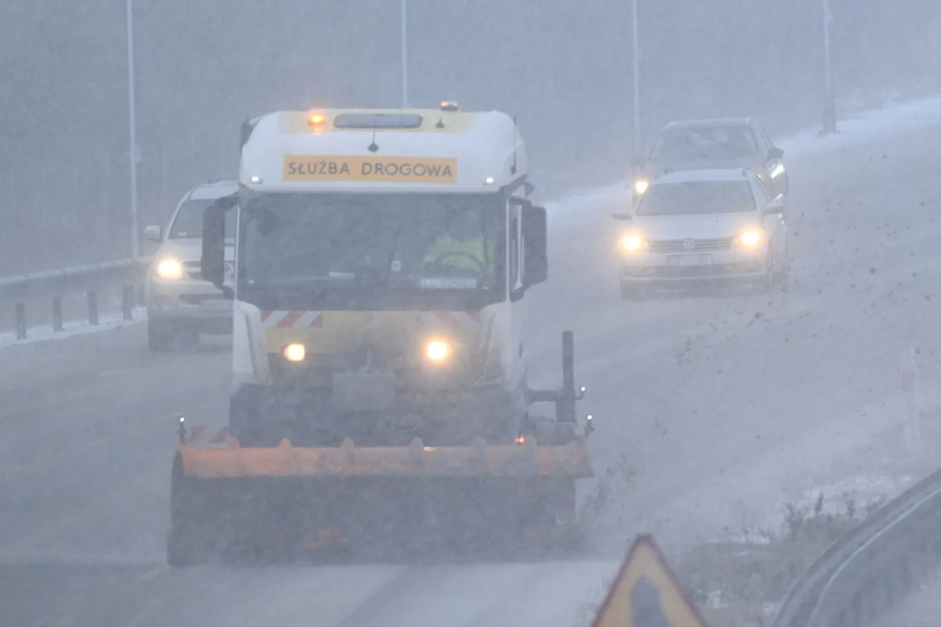 Zima nie odpuszcza. Jaka pogoda czeka nas w Sylwestra i na początku 2026 roku? Odśnieżarka z napisem 'Służba drogowa' pracuje na zaśnieżonej drodze w czasie gęstej śnieżycy, wokół niej jadą samochody z włączonymi światłami.