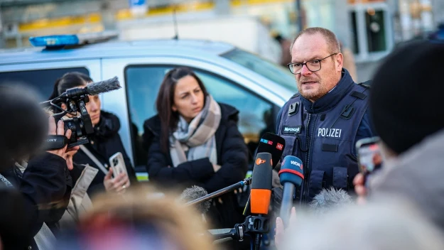 Policjant w kamizelce z napisem Polizei udziela wypowiedzi mediom, otoczony przez kilku dziennikarzy z mikrofonami i aparatami, w tle samochód policyjny oraz budynek o przeszklonych ścianach.