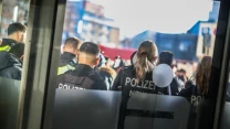 Grupa niemieckich policjantów stojących tyłem do obiektywu, ubranych w służbowe mundury, na tle zabudowy miejskiej w słoneczny dzień. Przednia część zdjęcia częściowo przysłonięta przez przeszklone drzwi z naklejonymi symbolami.