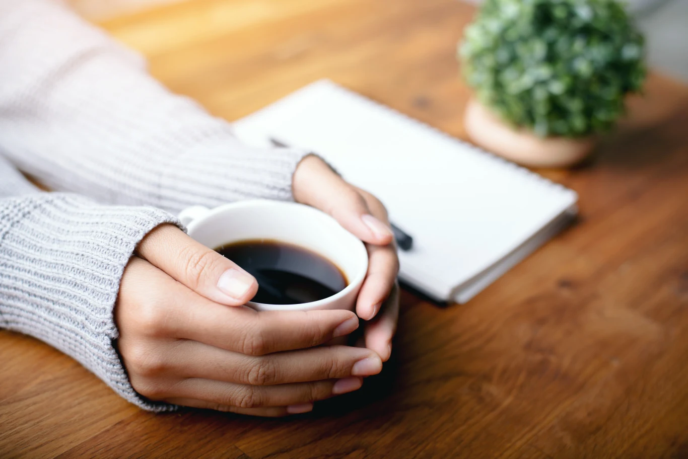 Dłonie trzymające filiżankę z czarną kawą, ubrane w szary sweter, na drewnianym stole obok notatnik i zielona roślina w doniczce.