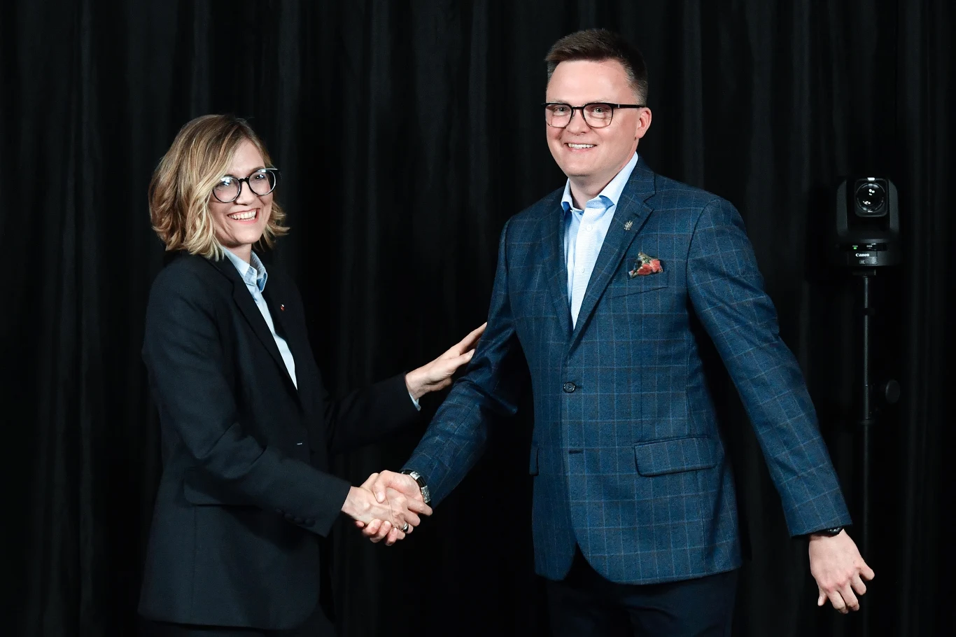 Magdalena Biejat i Szymon Hołownia podczas kampanii prezydenckiej Mężczyzna i kobieta ubrani w formalne garnitury podają sobie ręce, stojąc na tle czarnej kotary, oboje się uśmiechają.