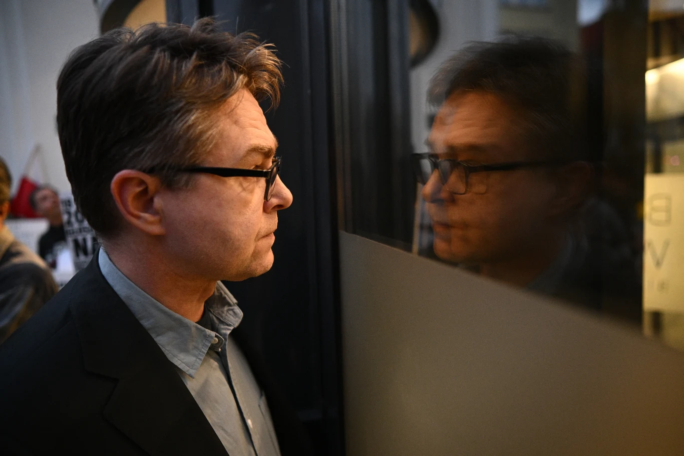 Mężczyzna w okularach stojący bokiem przy przezroczystej tafli, w której odbija się jego twarz, ujęcie w ciepłych barwach, w tle niewyraźne postacie i elementy architektury wnętrza.