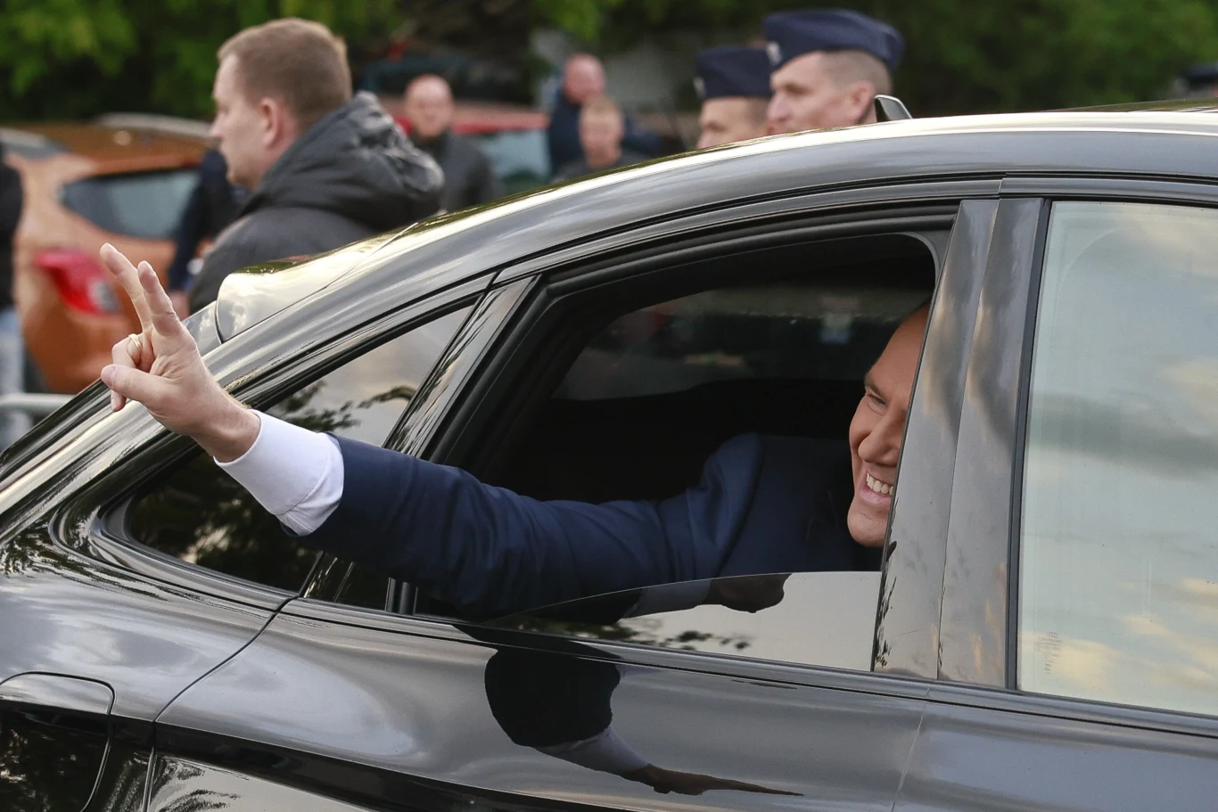 Karol Nawrocki nie był faworytem, a wygrał wybory prezydenckie Mężczyzna w garniturze pokazuje znak zwycięstwa przez opuszczoną szybę czarnego samochodu, uśmiechając się. W tle widoczne są inne osoby oraz kilka pojazdów.