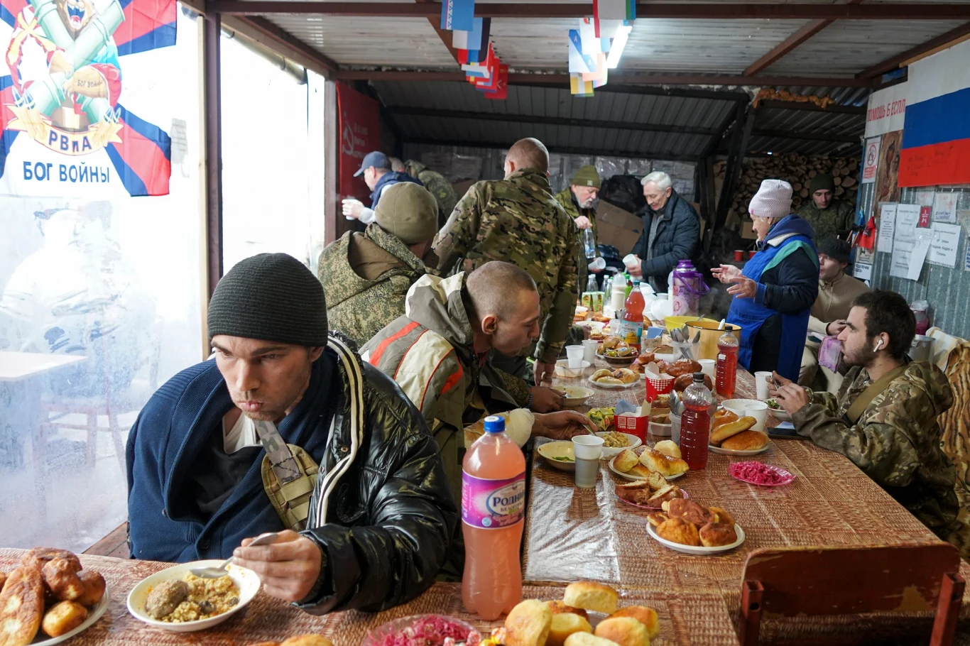 Żołnierze w mundurach siedzą przy stole zastawionym jedzeniem, spożywając posiłek w prowizorycznej stołówce udekorowanej rosyjskimi flagami oraz symbolami wojskowymi. Część osób jest zatopiona w rozmowach, inni skupieni na jedzeniu. W tle widać więcej ...