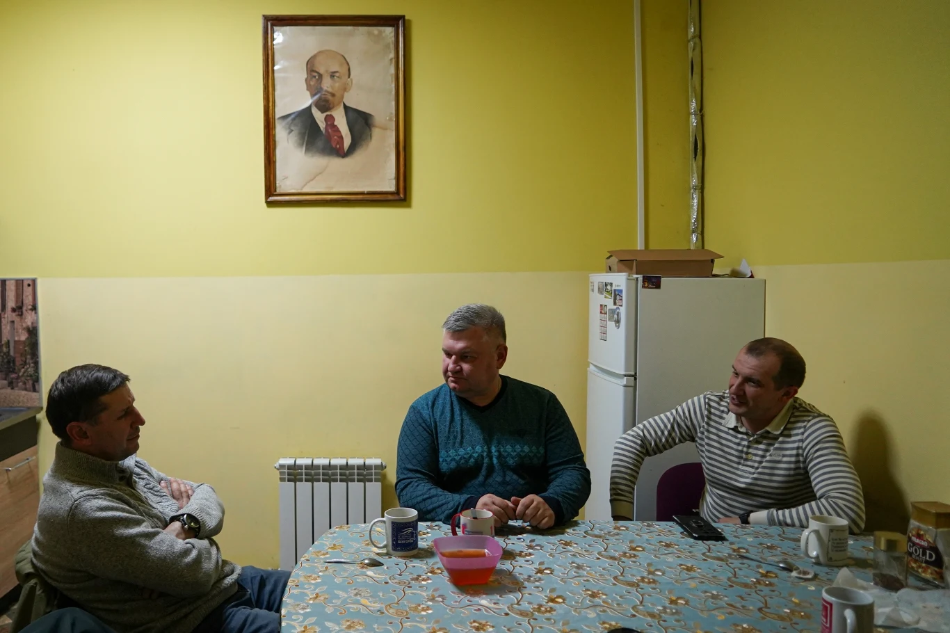 Trzech mężczyzn siedzi przy stole pokrytym obrusem we wzory w prostym pomieszczeniu o żółtych ścianach, nad nimi wisi oprawiony portret Włodzimierza Lenina, a za nimi znajduje się biały lodówka i kartony ustawione na górze.