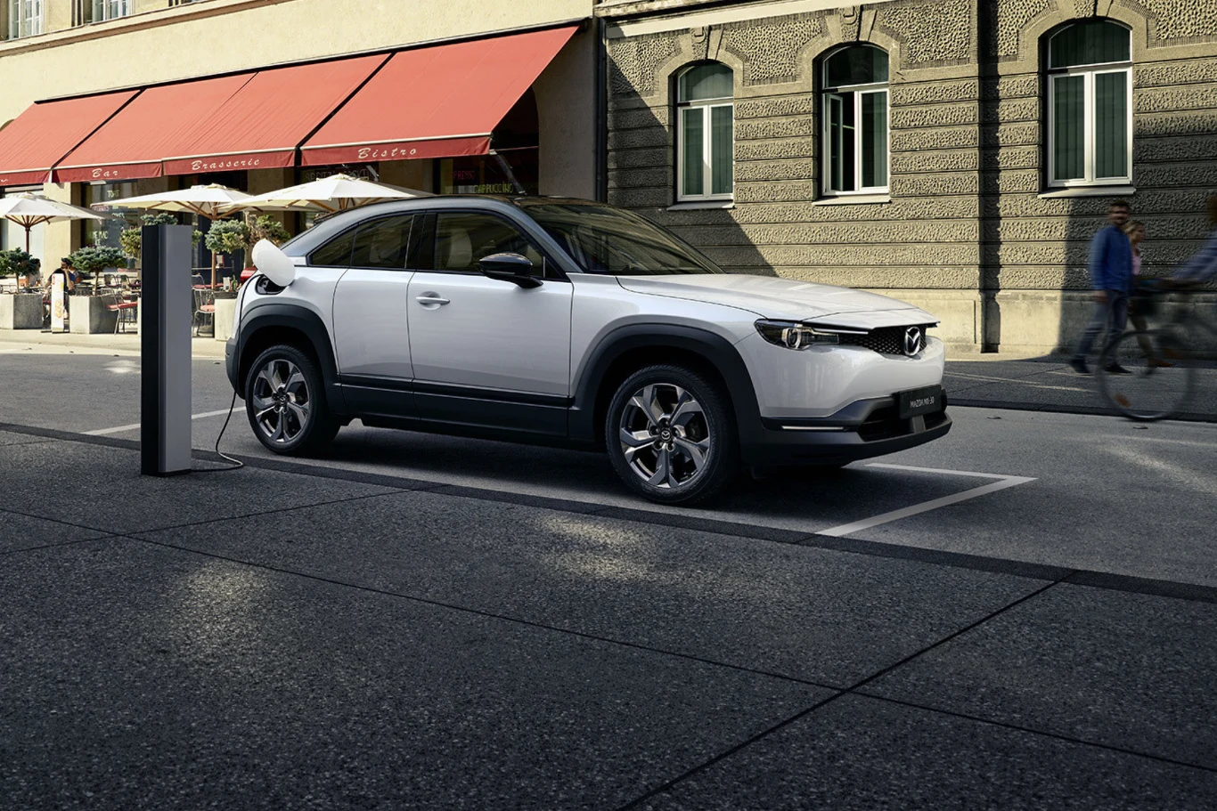 Elektryczny samochód marki Mazda ładowany przy miejskiej stacji ładowania, zaparkowany wzdłuż ulicy w otoczeniu miejskiej zabudowy, w tle widoczny rowerzysta oraz kawiarnia z czerwonym markizem.