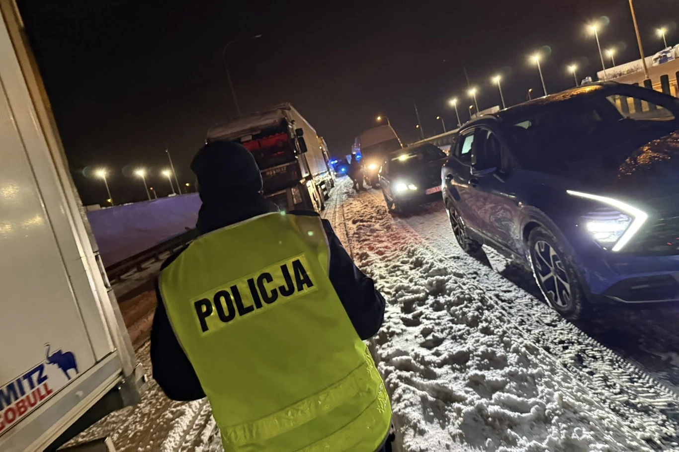 Zablokowana trasa S7. Ile paliwa zużywają stojące samochody? Policjant w żółtej kamizelce z napisem 'Policja' patroluje zaśnieżoną ulicę nocą, na której zatrzymane są samochody, w tym ciężarówka oraz auto osobowe, a w tle widoczne są latarnie i inne pojazdy.