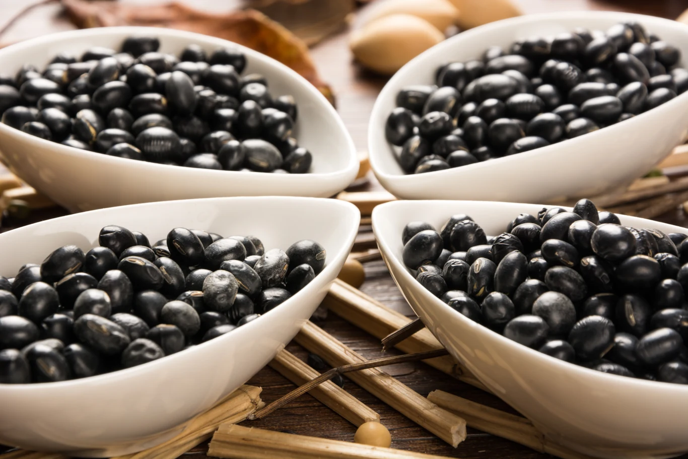 Cztery białe miseczki wypełnione czarną fasolą ustawione na drewnianych patyczkach, w tle niewyraźne elementy dekoracyjne.