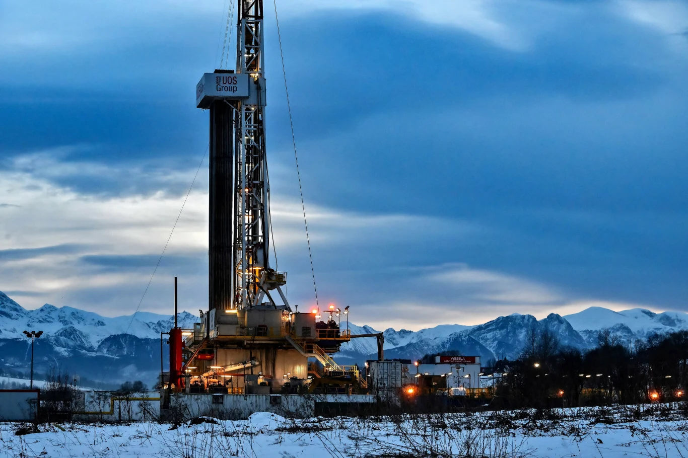 Odwiert geotermalny Bańska zakończono na początku 2025 roku Stacja wiertnicza otoczona śnieżnym krajobrazem z widokiem na górskie szczyty w tle. Konstrukcja główna jest oświetlona, a wokół znajdują się zabudowania techniczne. Nad całością rozpościera się wieczorne, pochmurne niebo.