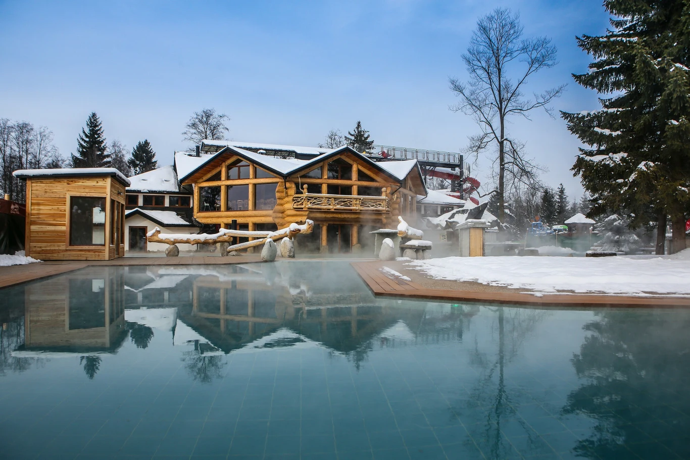 Na Podhalu odkryto głębsze struktury geotermalne, woda ma tam aż 100 st. C. Drewniany budynek w stylu góralskim otoczony śniegiem, przy którym znajduje się odkryty basen z parującą wodą. W tle rosną drzewa iglaste oraz kilka innych budynków, a wokół panuje zimowa aura.