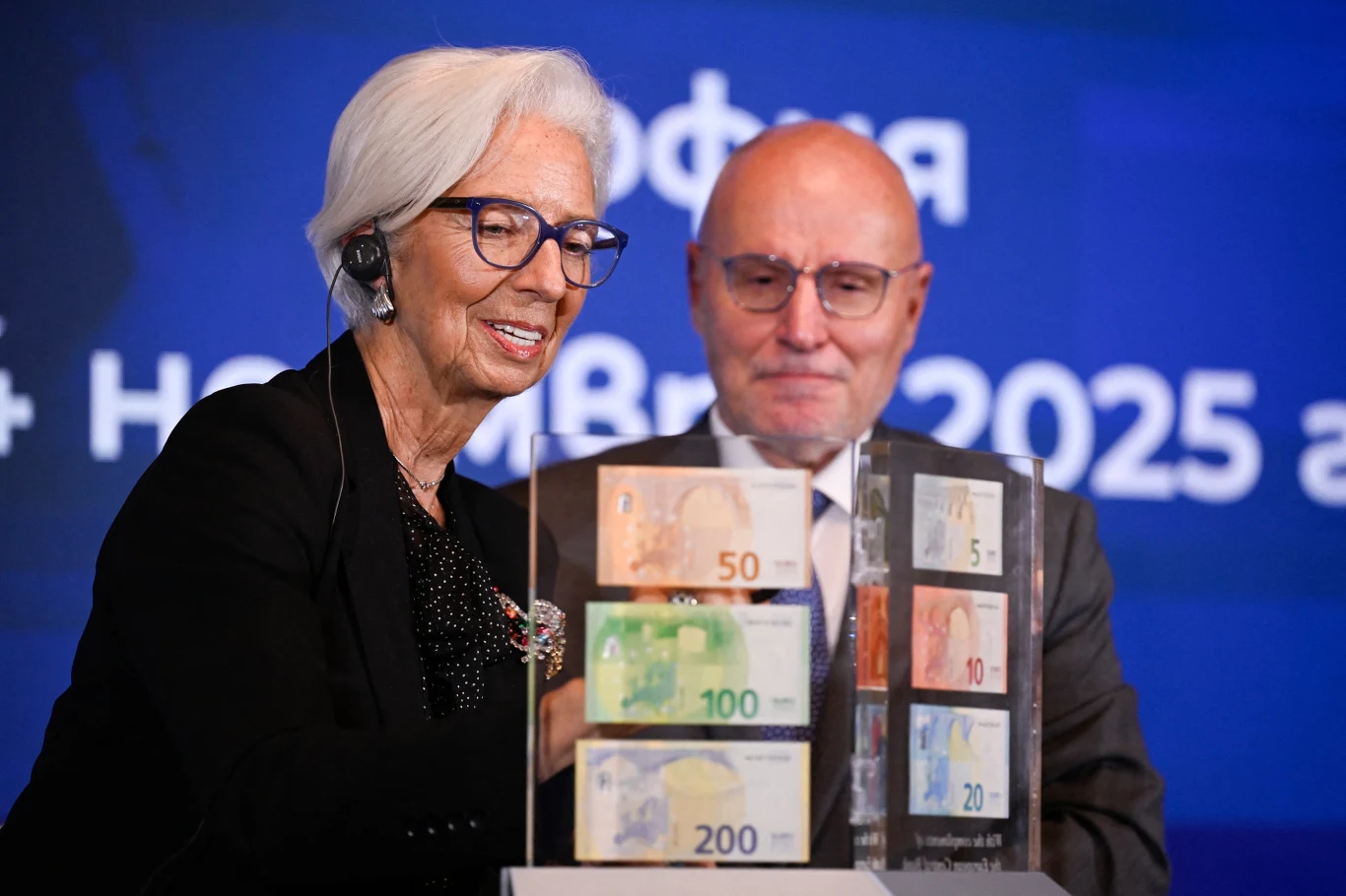 Starsza kobieta z białymi włosami i okularami oraz łysy mężczyzna w garniturze stoją przy przezroczystej gablocie z nowymi banknotami euro o nominałach 5, 10, 20, 50, 100 i 200, na tle niebieskiego ekranu z napisem.