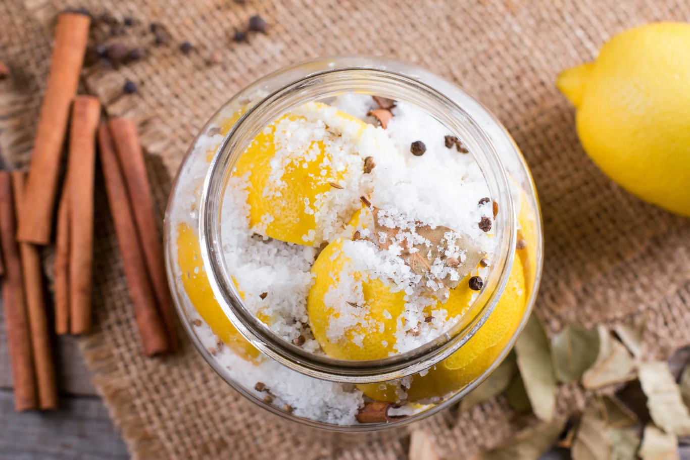 Pokrojone cytryny zasypane solą w szklanym słoju, obok laski cynamonu, liście laurowe i cała cytryna na jutowym materiale.