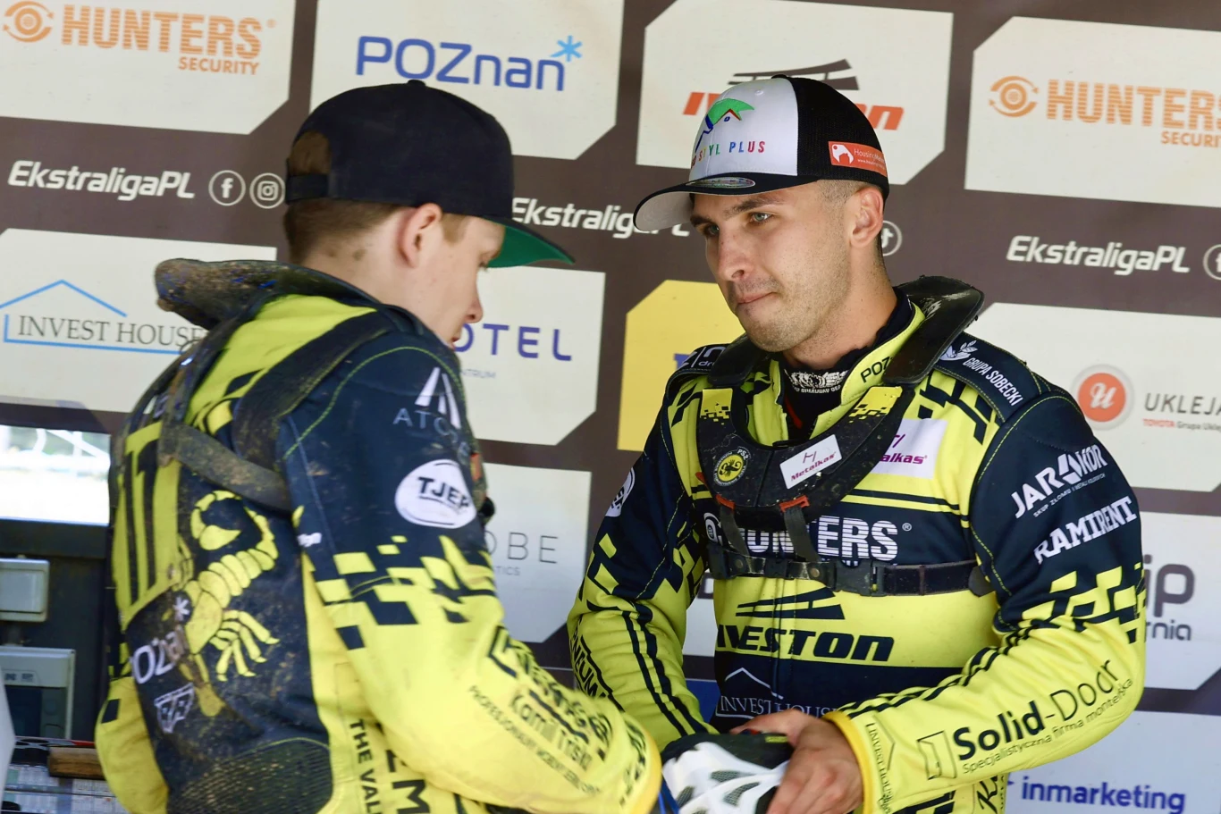 Szymon Szlauderbach z Francisem Gustsem Dwóch żużlowców w żółto-czarnych strojach drużynowych ściska sobie dłonie, stojąc na tle tablicy z logotypami sponsorów i organizatorów zawodów.