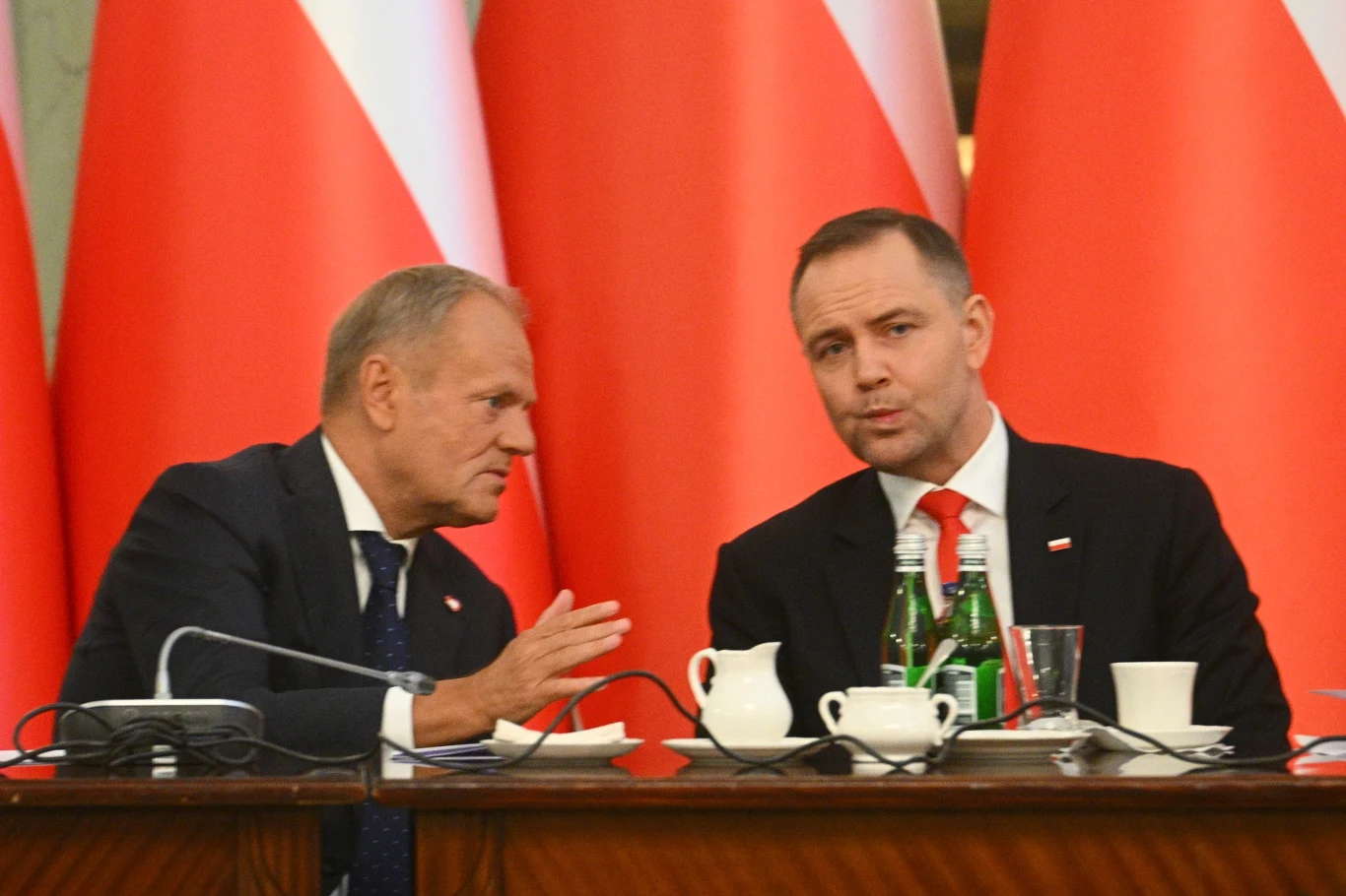 Dwóch mężczyzn w garniturach siedzi przy stole, rozmawiają ze sobą. W tle widoczne duże biało-czerwone flagi. Na stole znajdują się butelki z wodą, filiżanki i dzbanek.