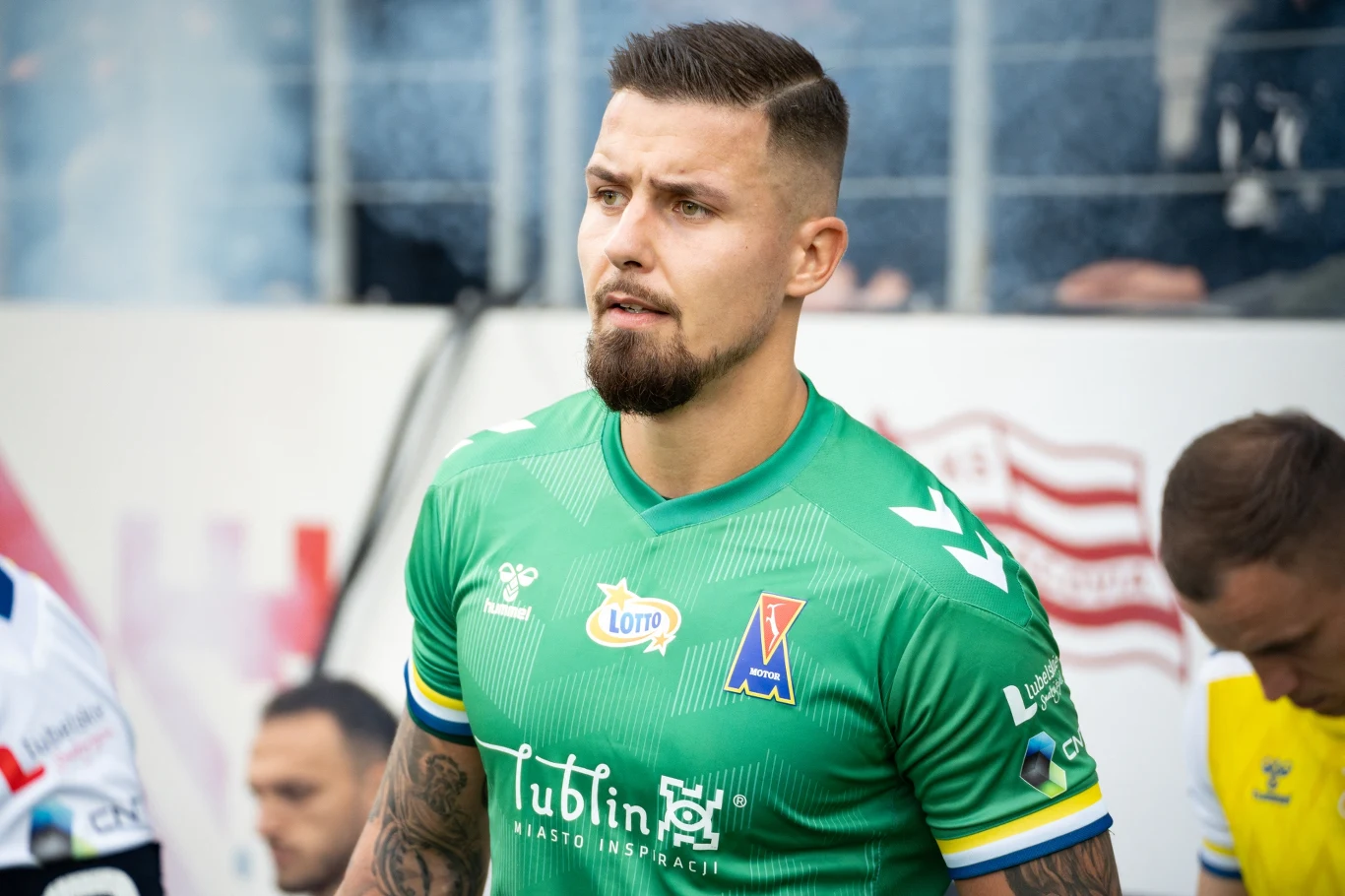 Mężczyzna w zielonej koszulce sportowej z logotypami sponsorów stoi na tle boiska oraz innych zawodników, skupiony i gotowy do gry.