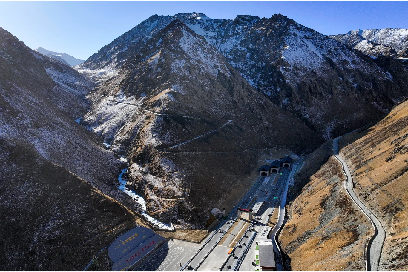 Górska dolina z wysokimi, częściowo ośnieżonymi szczytami, przez którą przebiega nowoczesna droga w tunelu. Wjazd do tuneli znajduje się u podnóża stromych zboczy, a wzdłuż drogi widoczne są pojazdy i infrastruktura drogowa. Widok obejmuje również zakr...