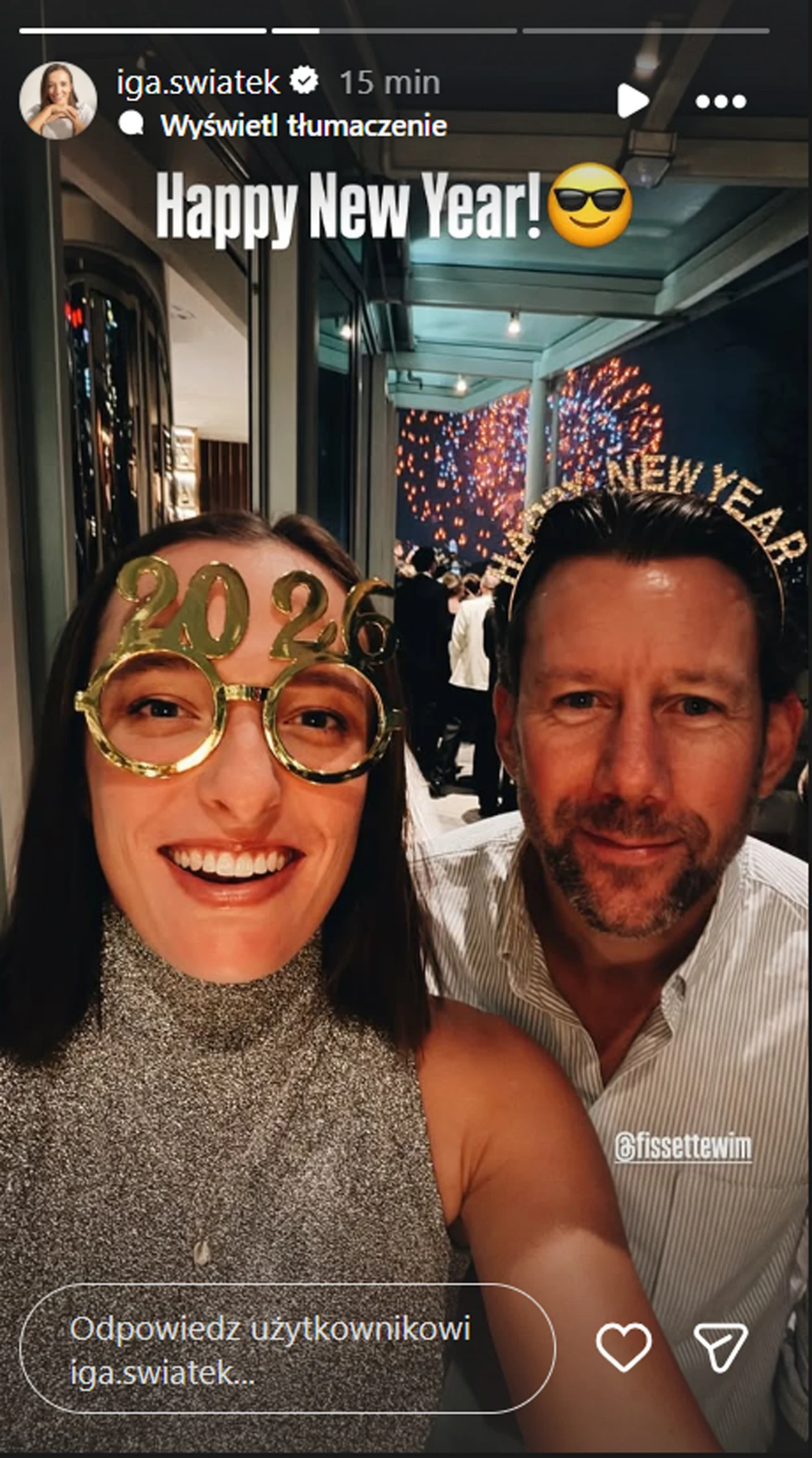 Dwie uśmiechnięte osoby pozują do selfie w środku pomieszczenia. Jedna z nich nosi okulary z napisem 2024, a w tle widoczne są ozdoby i napis Happy New Year wraz ze sztucznymi ogniami.