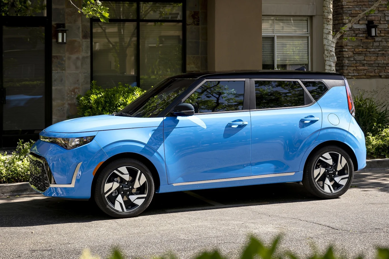 Nowoczesny, niebieski samochód osobowy typu crossover zaparkowany na ulicy, w tle budynek mieszkalny z dużym oknem i elementami kamiennej elewacji, wokół zieleń i drzewa rzucające cień na pojazd.