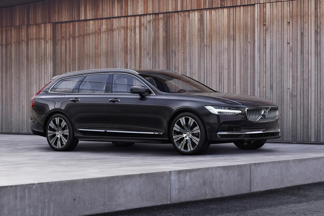 Luksusowe czarne kombi Volvo z chromowanymi detalami, osadzone na dużych alufelgach, stoi na tle drewnianej ściany, podkreślając nowoczesny design i elegancję auta.