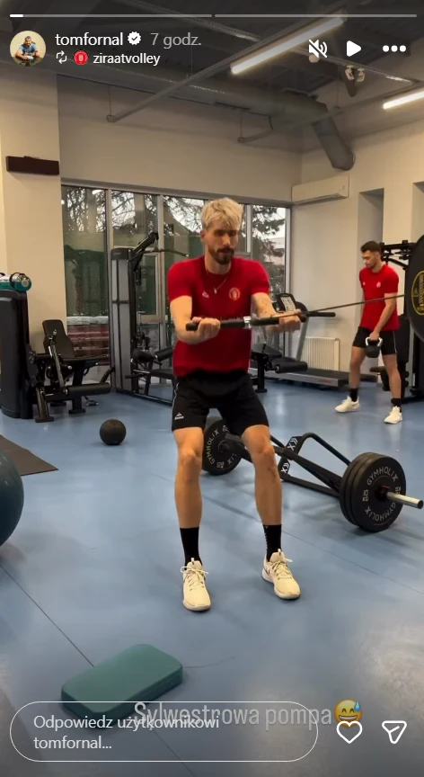 Dwóch mężczyzn w sportowych strojach ćwiczy na siłowni, wykonując ćwiczenia przy użyciu różnych sprzętów do treningu siłowego, w tle widoczne są maszyny i ciężary.