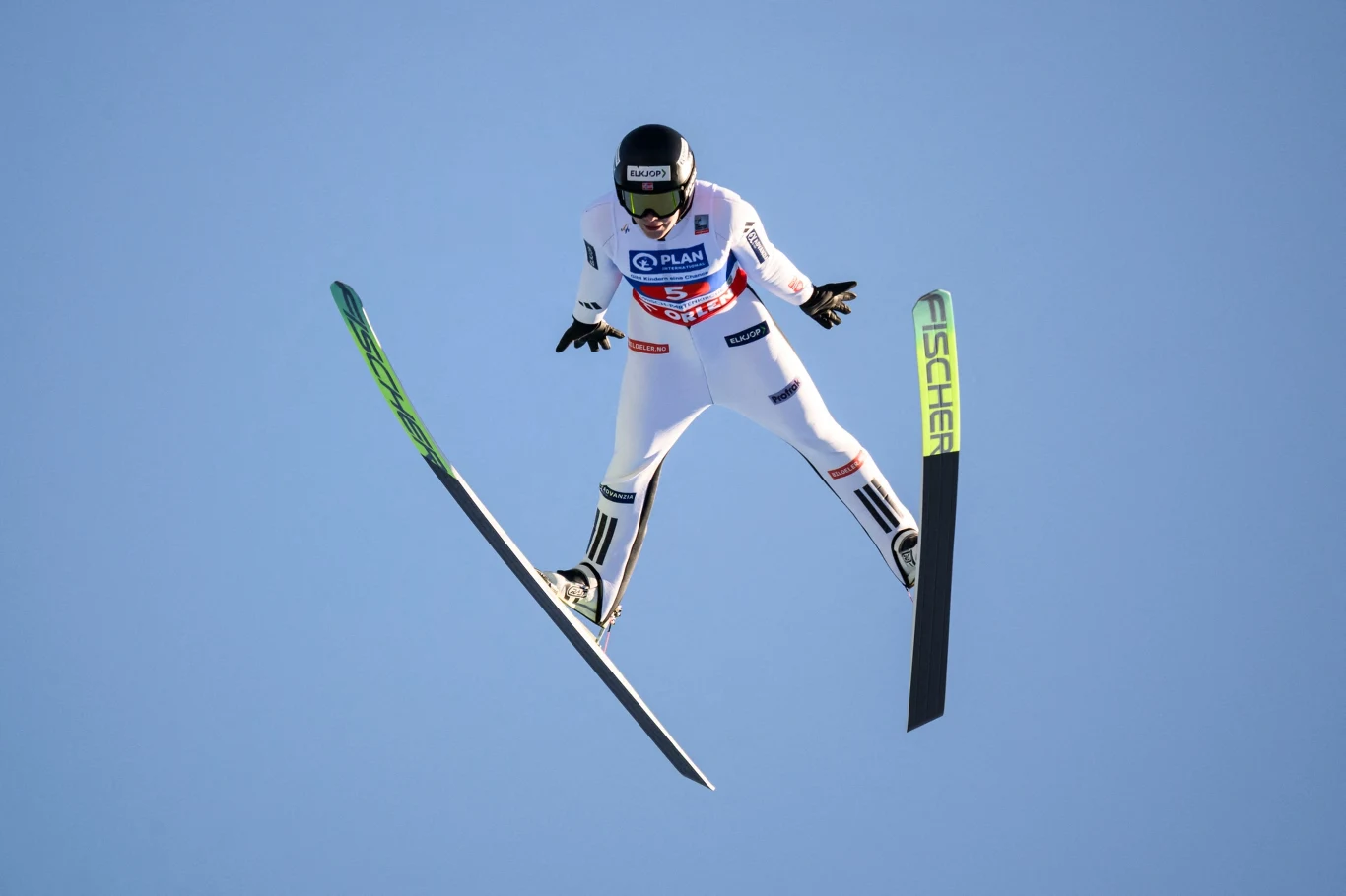 Skoczek narciarski w locie podczas konkursu skoków narciarskich, sylwetka sportowca w białym kombinezonie na tle błękitnego nieba, narty rozstawione szeroko, na nartach widoczne logo producenta.