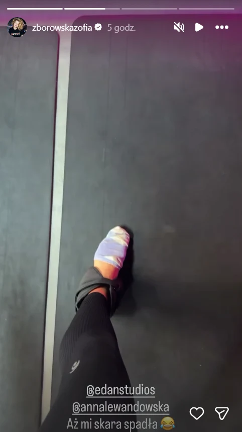 Lewą stopę w czarnej legginsie oraz sportowym obuwiu z widoczną częściowo zsuniętą kolorową skarpetką na ciemnej podłodze prawdopodobnie w siłowni lub studiu treningowym.