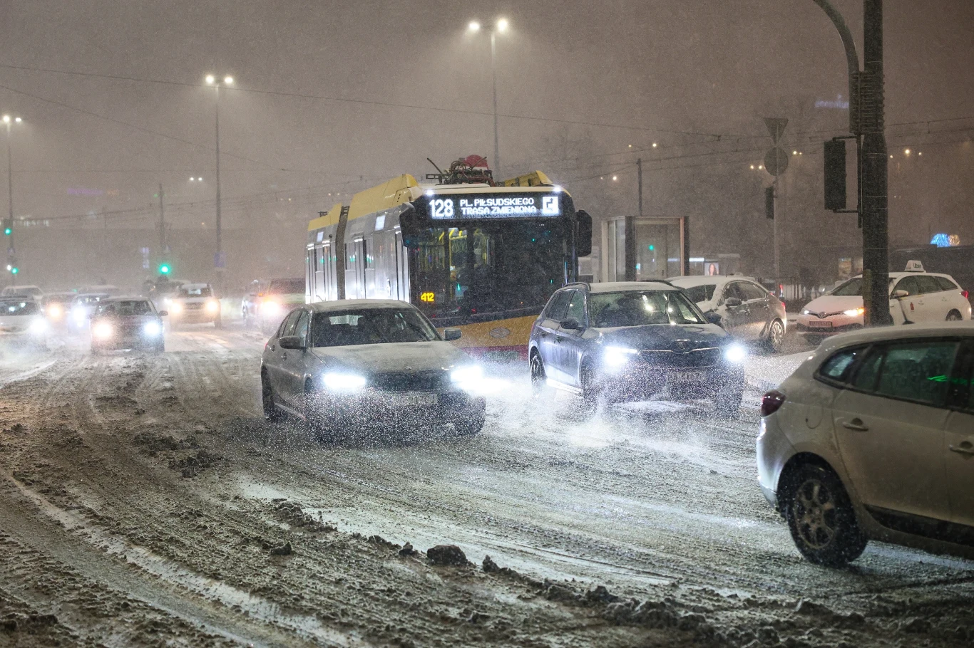 Pogoda w Polsce nie rozpieszcza. Zima trzyma mocno, wydano nowe ostrzeżenia Samochody oraz trolejbus poruszają się po zaśnieżonej, ruchliwej ulicy w centrum miasta nocą, światła pojazdów rozświetlają zamieć śnieżną, a intensywne opady powodują utrudnienia w ruchu.