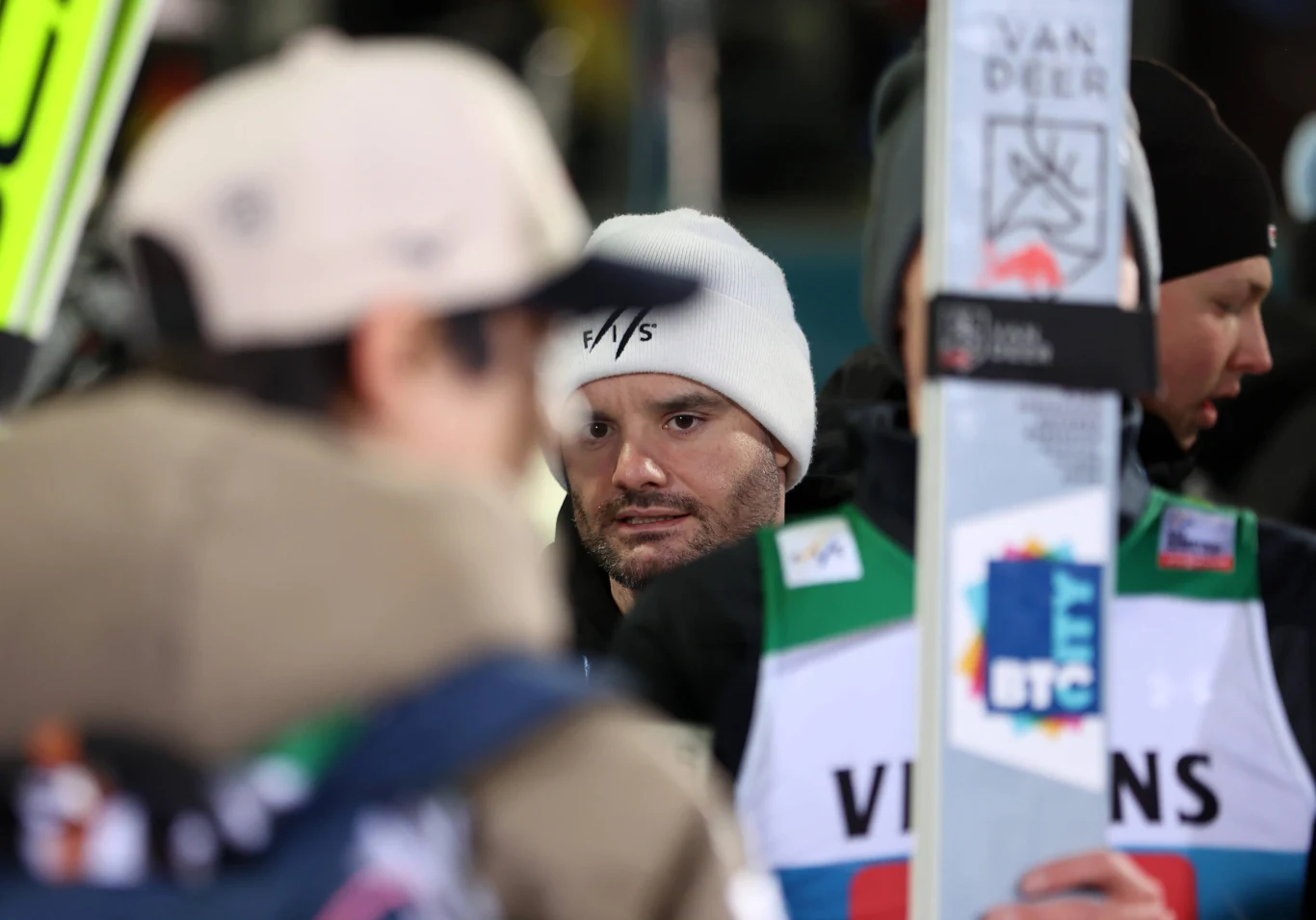 Skoczek narciarski w białej czapce z napisem F/IS w otoczeniu innych zawodników na zawodach, w tle widoczna tablica z napisem Van Deer i kolorowa naszywka BTC na kamizelce.