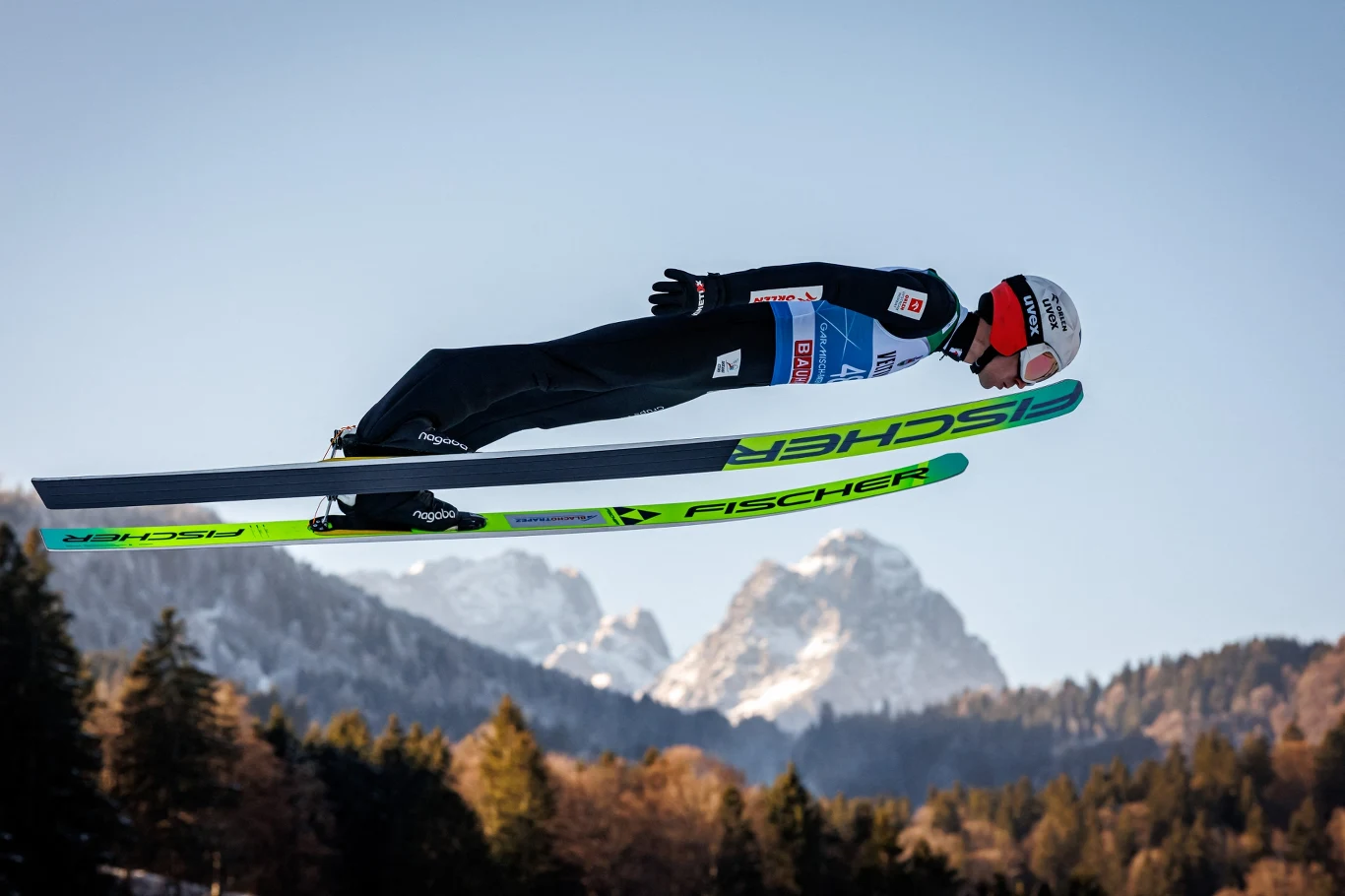 Skoczek narciarski w locie nad śnieżnymi górami, w tle drzewa oraz ośnieżone szczyty, zawodnik w sportowym kombinezonie i kasku, dynamiczna sylwetka podkreślająca ruch i prędkość.
