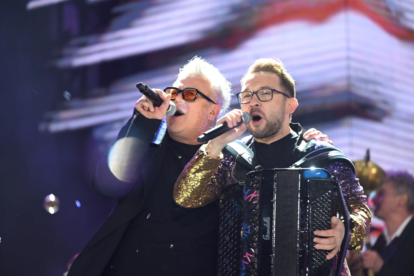 Dwóch śpiewających mężczyzn obejmuje się na scenie podczas występu. Jeden z nich gra na akordeonie, obaj trzymają mikrofony. Na pierwszym planie widoczny jest efektowny strój ze złotymi rękawami oraz kolorowe światła sceniczne.