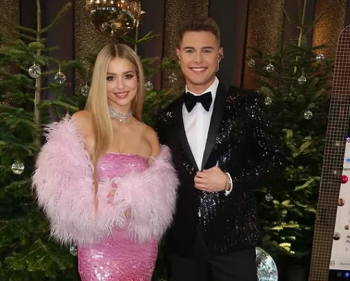 Elegancko ubrana młoda para pozuje na tle ozdobionych świątecznie choinek, kobieta w różowej cekinowej sukni z futerkowym narzutką, mężczyzna w czarnym garniturze z muchą i błyszczącą marynarką.