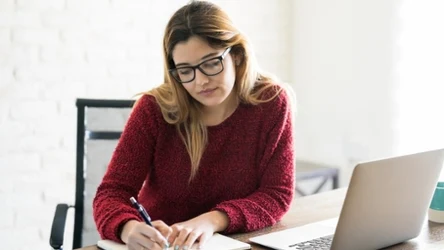 Młoda kobieta w bordowym swetrze, robi notatki przy biurku