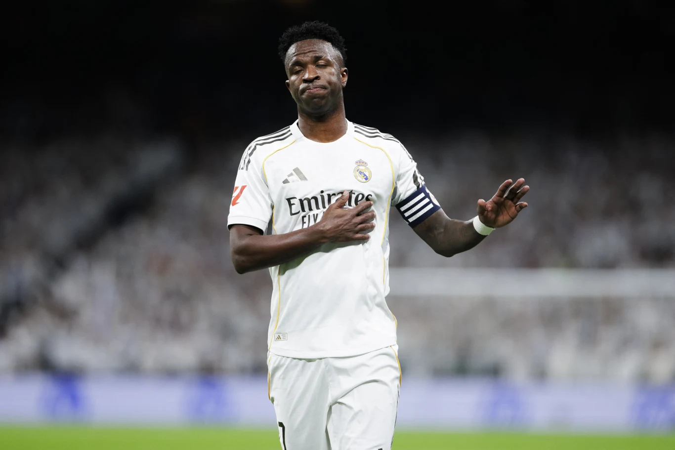 Piłkarz w stroju Realu Madryt z opaską kapitana na ramieniu, z wyrazem niezadowolenia na twarzy i gestem uspokajającym, na tle rozmytej widowni stadionu.