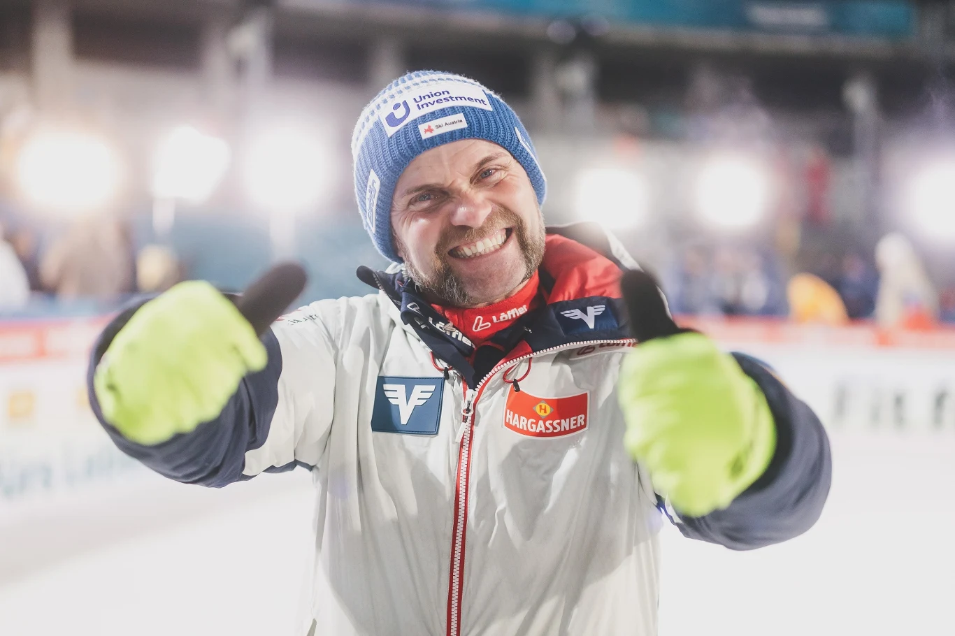 Uśmiechnięty mężczyzna w sportowej kurtce oraz czapce z napisem stoi w jasnym świetle, pokazując kciuki do góry w obu dłoniach. W tle rozmyte światła oraz niewyraźne postaci.