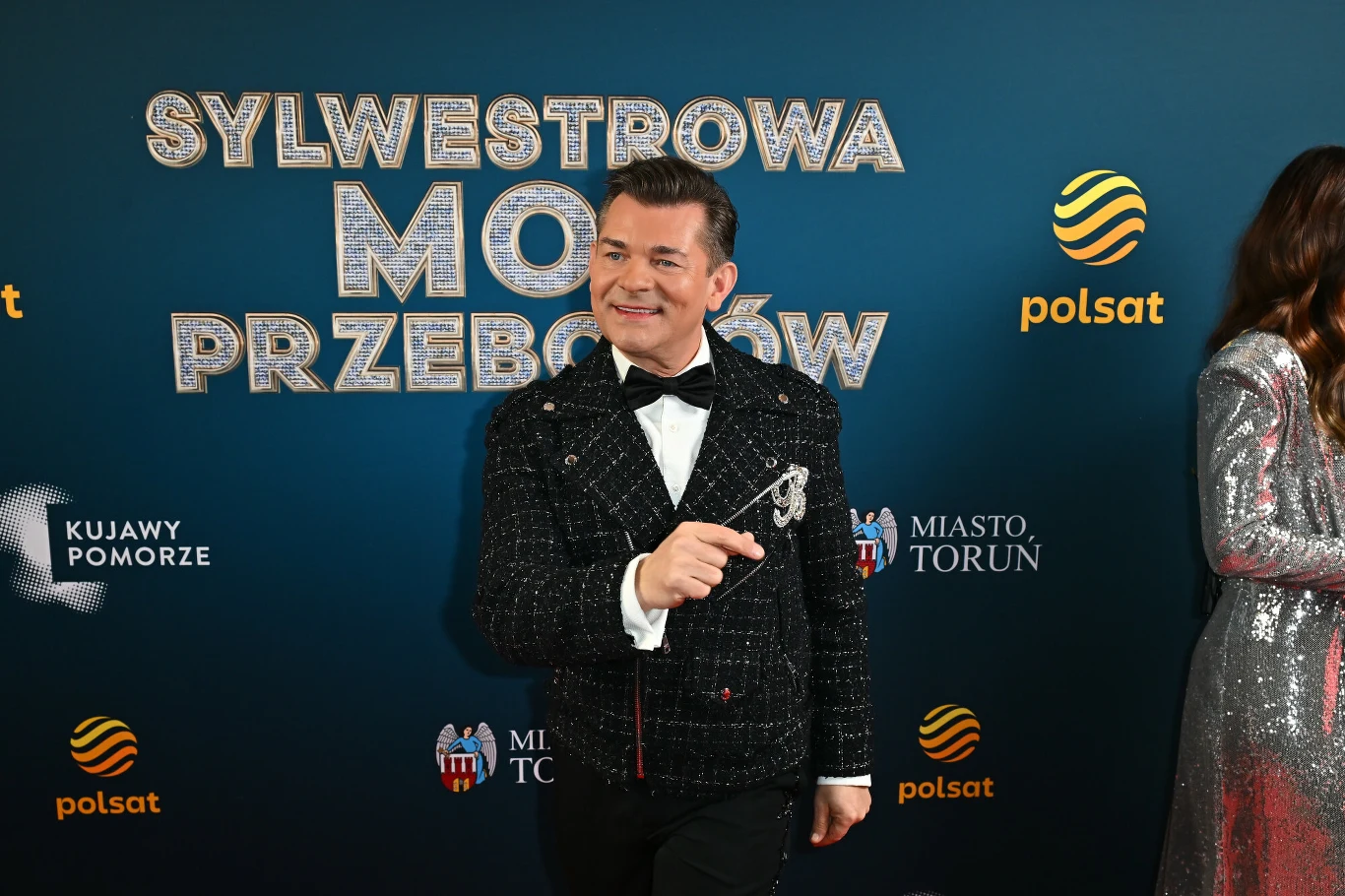 Zenek Martyniuk był jedną z gwiazd Sylwestrowej Mocy Przebojów Elegancki mężczyzna w ciemnej marynarce, białej koszuli i muchą stoi na tle ścianki z logo Polsat i napisem Sylwestrowa Moc Przebojów, obok znajduje się fragment drugiej osoby w srebrnej kreacji.