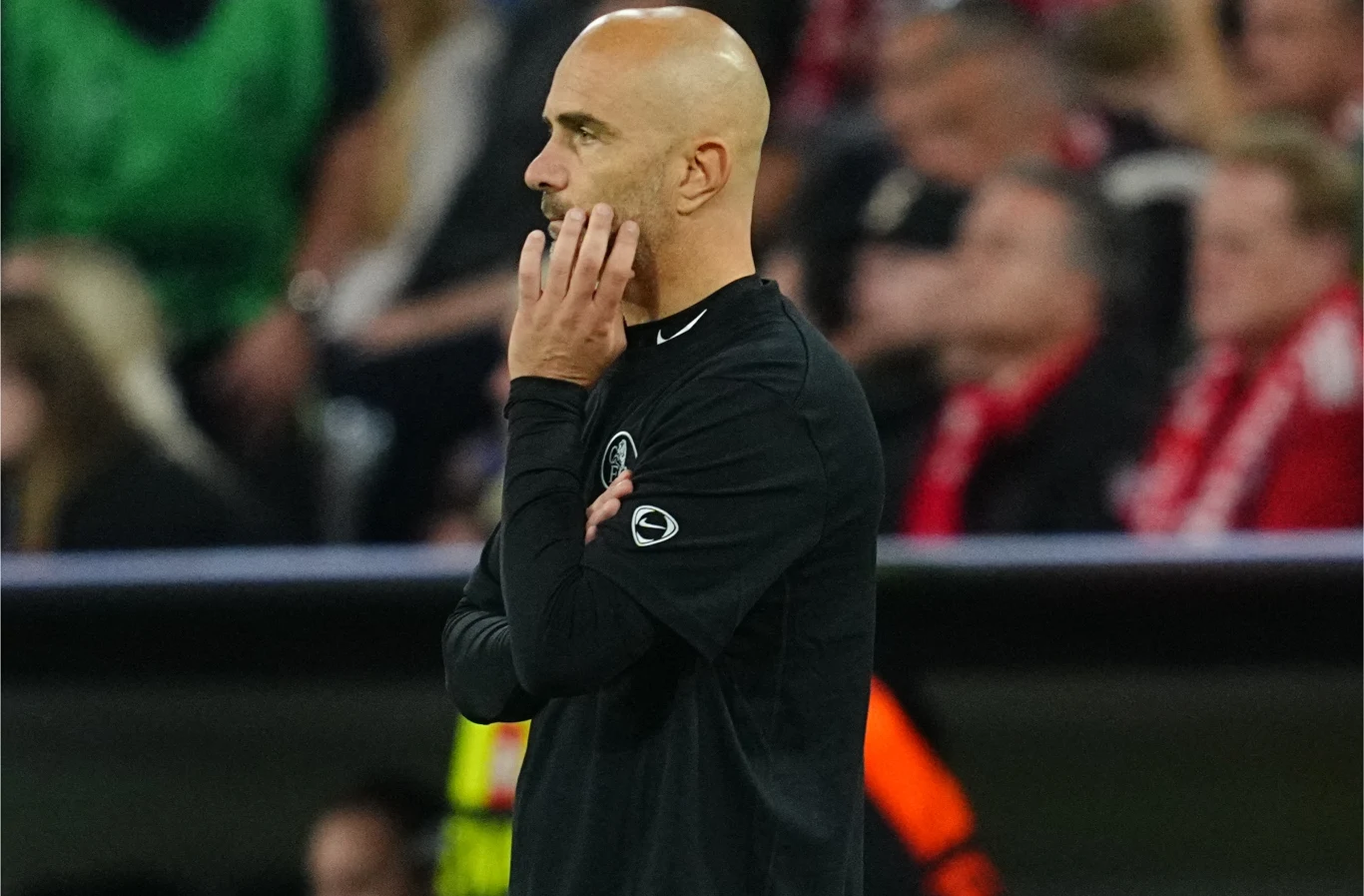 Enzo Maresca Łysy mężczyzna w ciemnym ubraniu stoi bokiem z ręką opartą o twarz, w tle rozmyte sylwetki ludzi na trybunach.