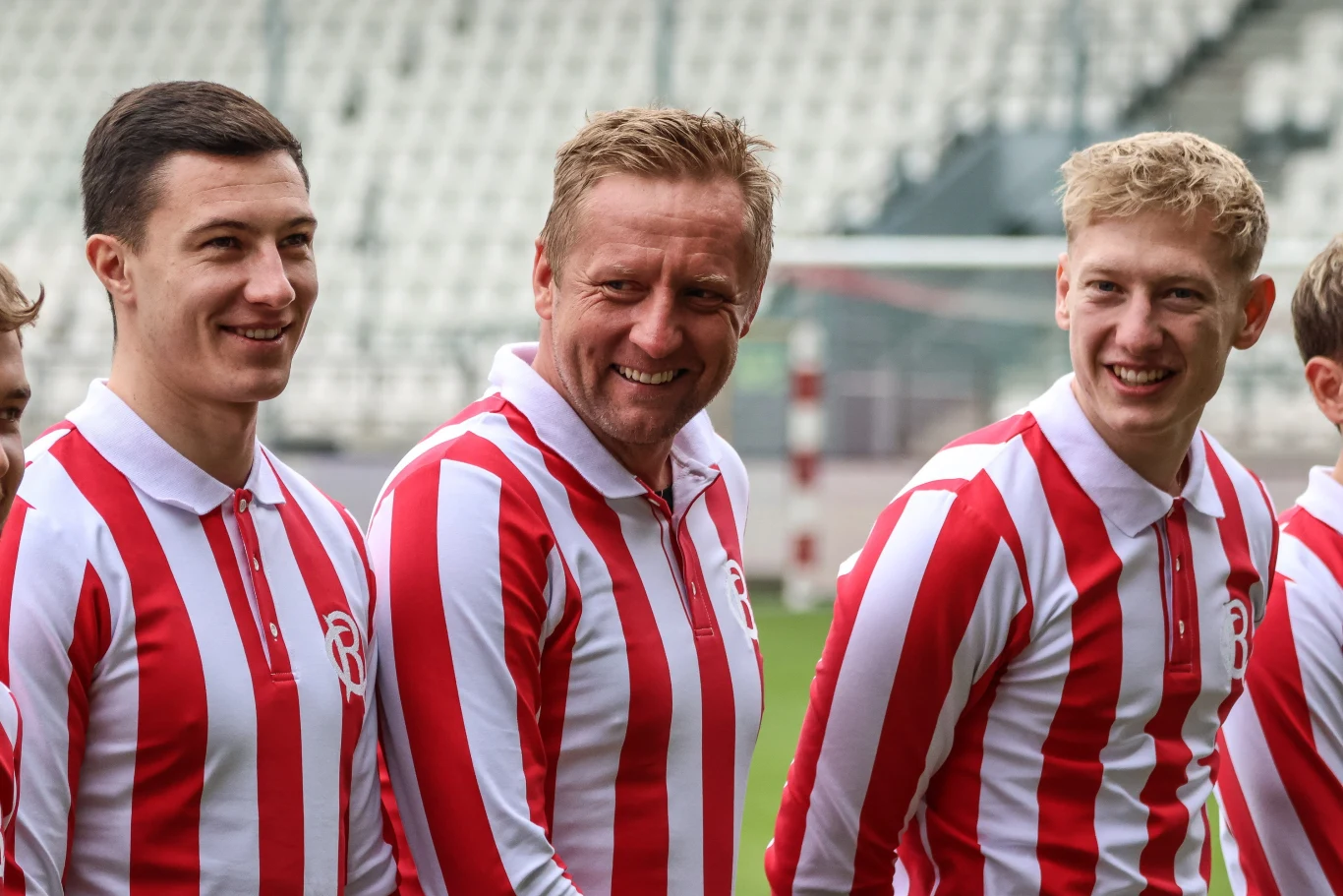 Trzech uśmiechniętych mężczyzn w biało-czerwonych pasiastych koszulkach sportowych stoi na stadionie piłkarskim, w tle widać puste trybuny.