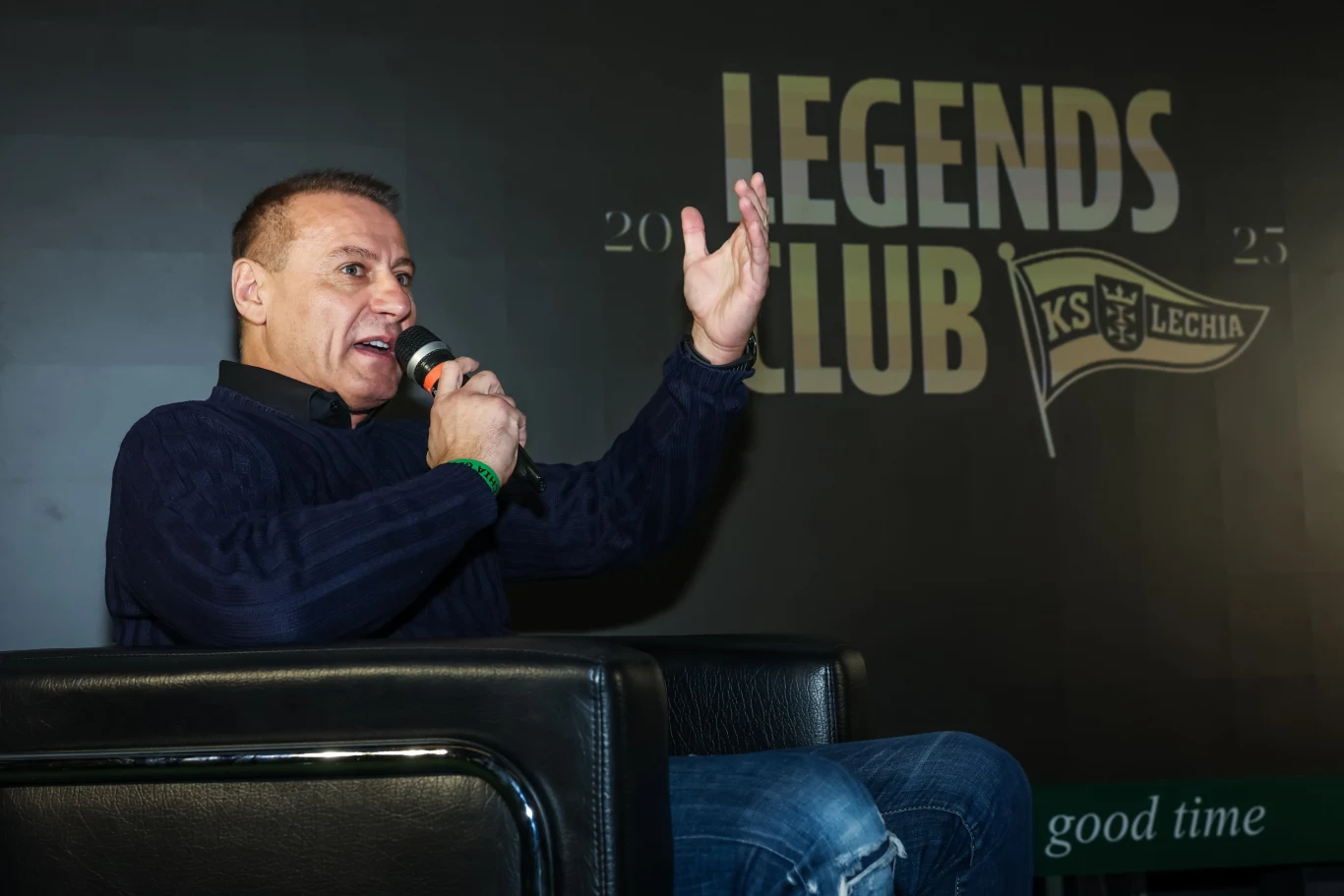 Mężczyzna siedzi na czarnym fotelu i przemawia do mikrofonu, gestykulując jedną ręką. W tle widoczny wyraźny napis LEGENDS CLUB oraz logo KS Lechia.