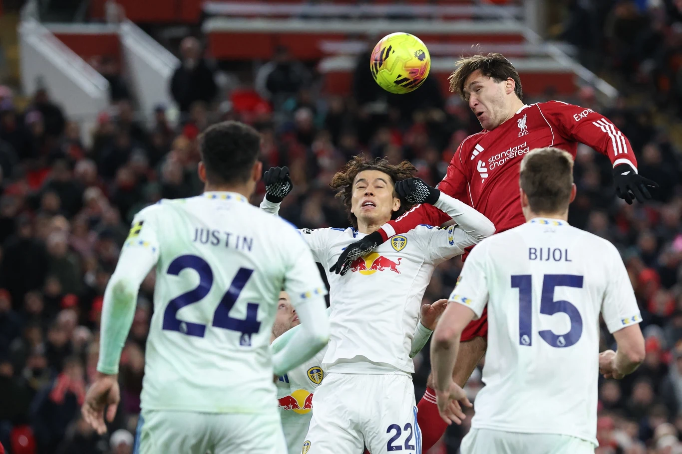 Piłkarz w czerwonym stroju wyskakuje wysoko do piłki, rywalizując w powietrznym pojedynku z zawodnikami drużyny w białych koszulkach na tle trybun pełnych kibiców.